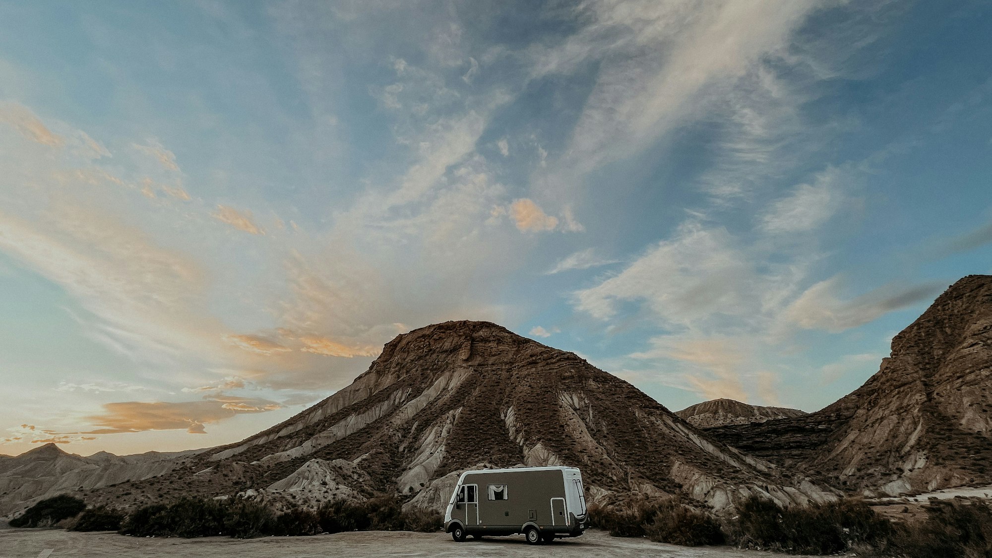 Ein Wohnmobil steht vor einer kahlen Hügelkette, die Wolken im Hintergrund scheinen gelblich.