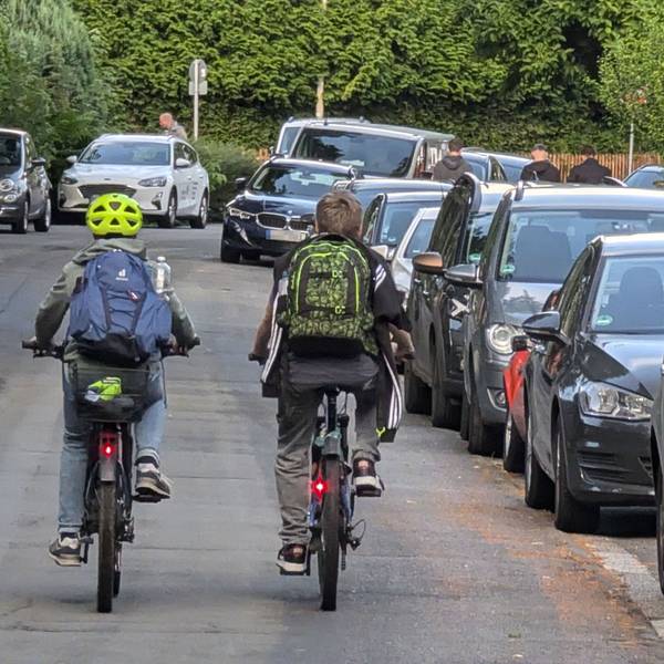 Zwei Kinder mit Rucksäcken auf dem Rücken fahren nebeneinander mit Fahrrädern über die Bonnstraße.