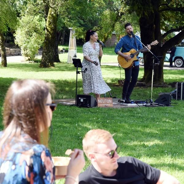 Zwei Musiker spielen und singen auf einer grünen Wiese, auf der Menschen picknicken.