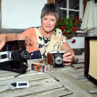 Eine Frau sitzt an einem Gartentisch und hält eine Hand über einen Diaprojektor. Auf dem Tisch liegen außerdem Dias, eine Leinwand und eine alte Fotokamera.