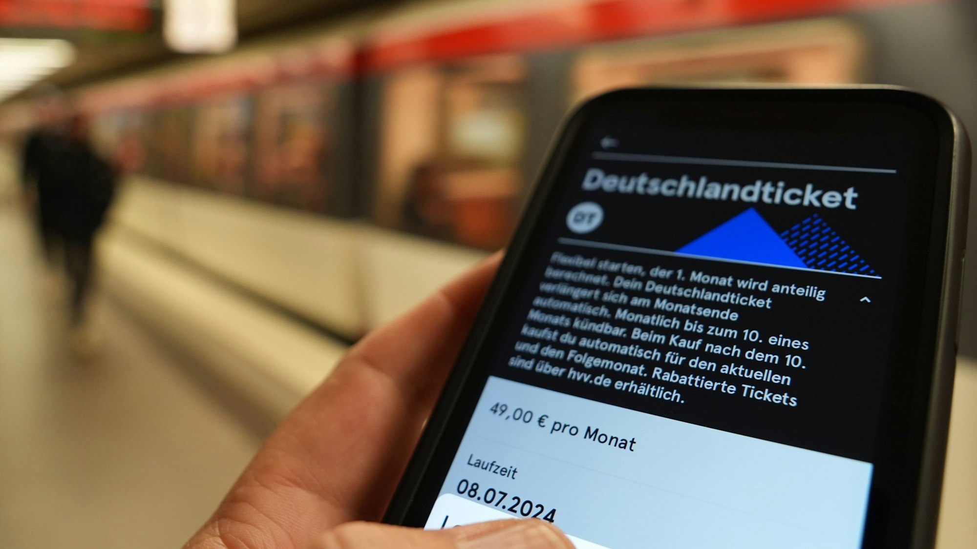 Ein Fahrgast hält ein Smartphone mit einem digitalen Deutschlandticket an einer U-Bahnstation in der Hand.