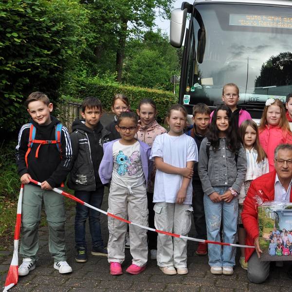 Grundschulkinder aus Golbach beim Abschiedsfoto mit Borislav Radovic. Sie stehen hinter einem Flatterband vor dem Schulbus, Radovic kniet bei den Kindern und hält eine Fotografie in Händen.