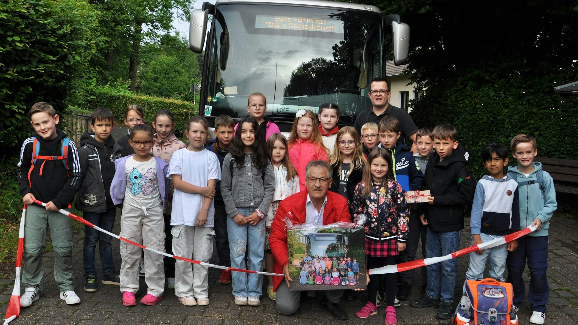 Grundschulkinder aus Golbach beim Abschiedsfoto mit Borislav Radovic. Sie stehen hinter einem Flatterband vor dem Schulbus, Radovic kniet bei den Kindern und hält eine Fotografie in Händen.