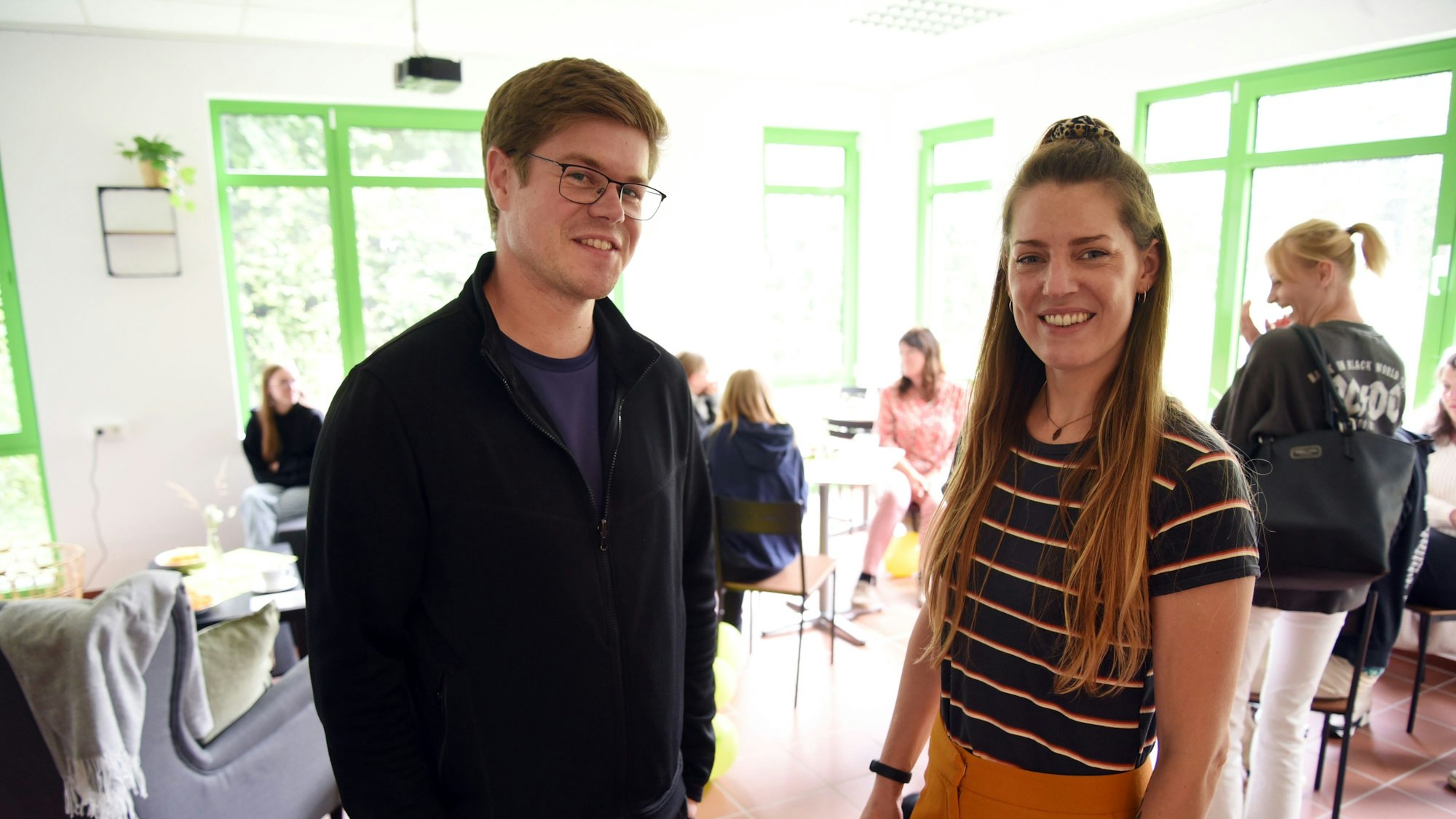 Ein Mann und eine Frau stehen nebeneinander in einem hellen Raum, im Hintergrund sitzen und stehen mehrere Menschen.
