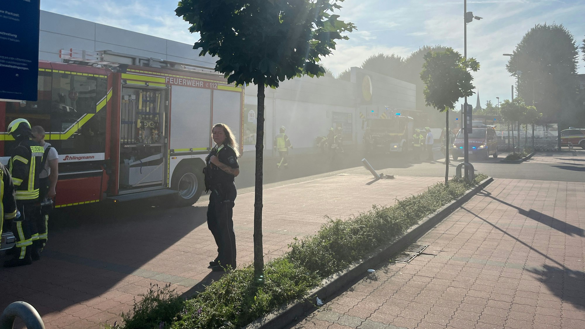 Rauch zieht über einen Parkplatz, auf dem Feuerwehrfahrzeuge stehen.