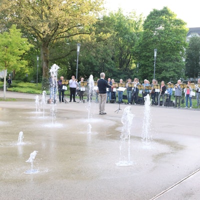 Ein Blasorchester spielt, im Vordergrund sieht man ein Wasserspiel.