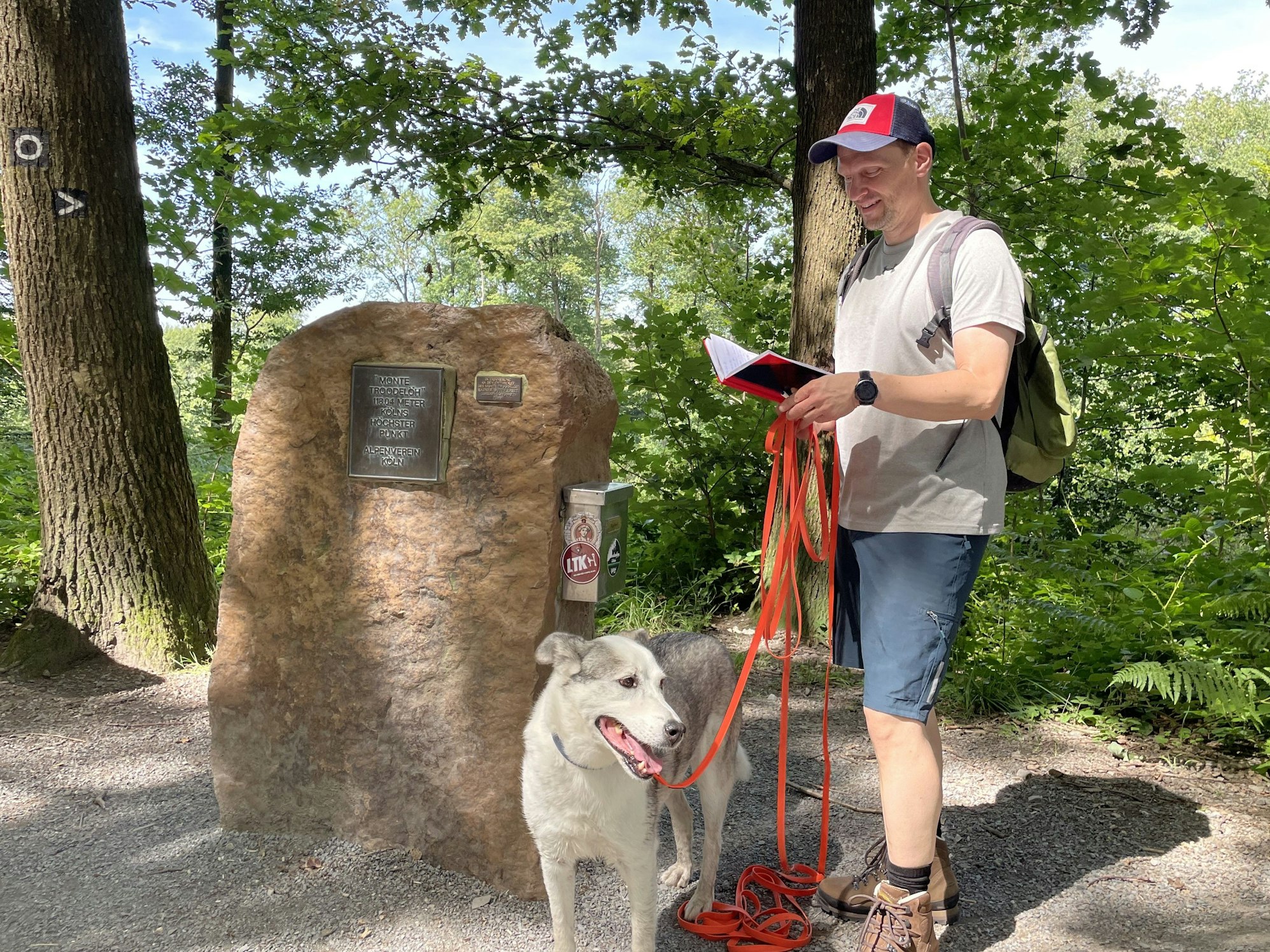 Ein Mann mit Hund steht vor dem Gipfelstein am Monte Troodelöh, dem höchsten Berg Kölns.