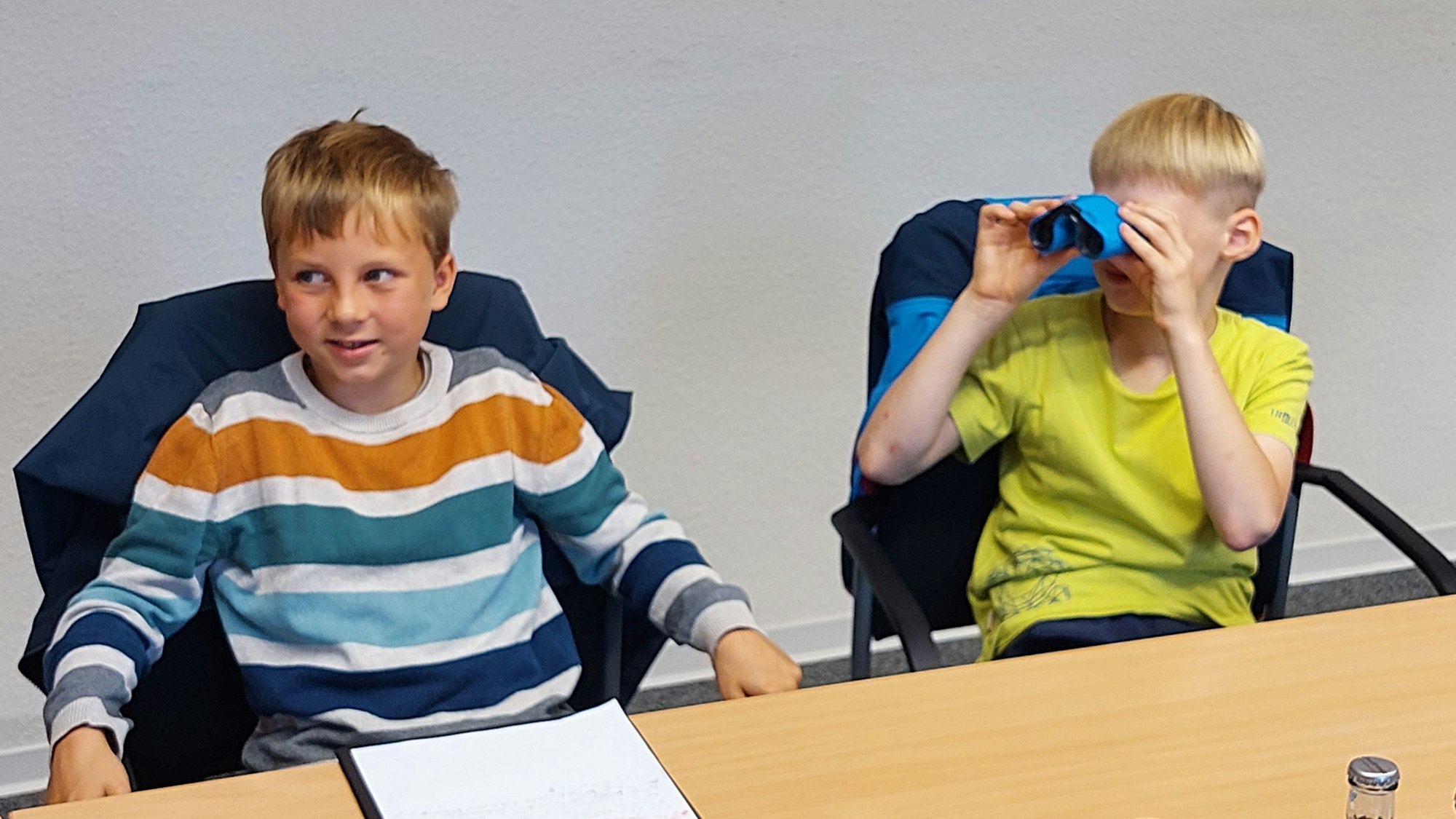 Zwei Jungen sitzen auf Stühlen an einem Tisch. Einer guckt durch ein Fernglas.