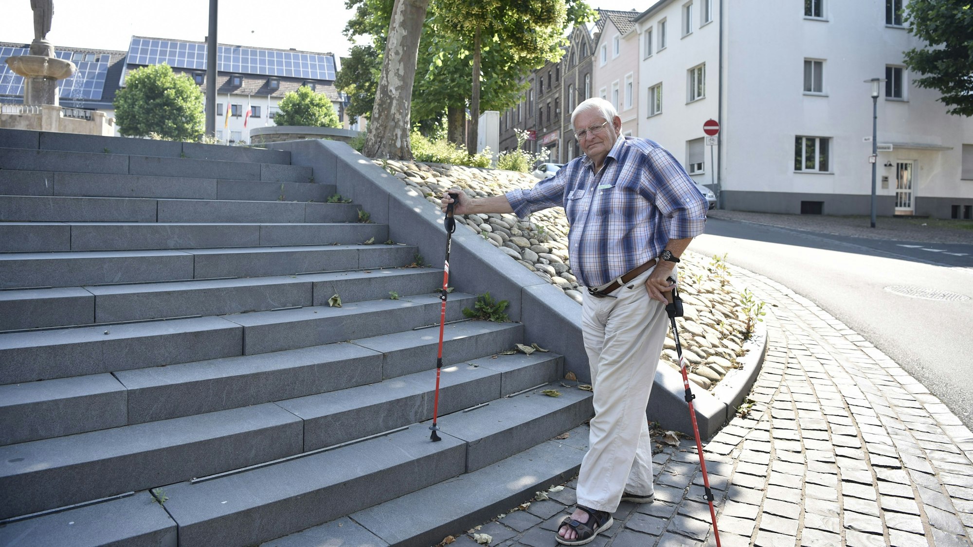 Wolfgang Lüttgen steht an einer Treppe, die kein Geländer hat. Er hat in jeder Hand einen Stock und trägt eine helle, lange Hose, ein kariertes Hemd mit hochgekrempelten Ärmeln sowie Sandalen.