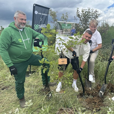 Lukas Podolski und weitere Männer und eine Frau stehen in einem Wald in Lindlar und pflanzen einen Baum.