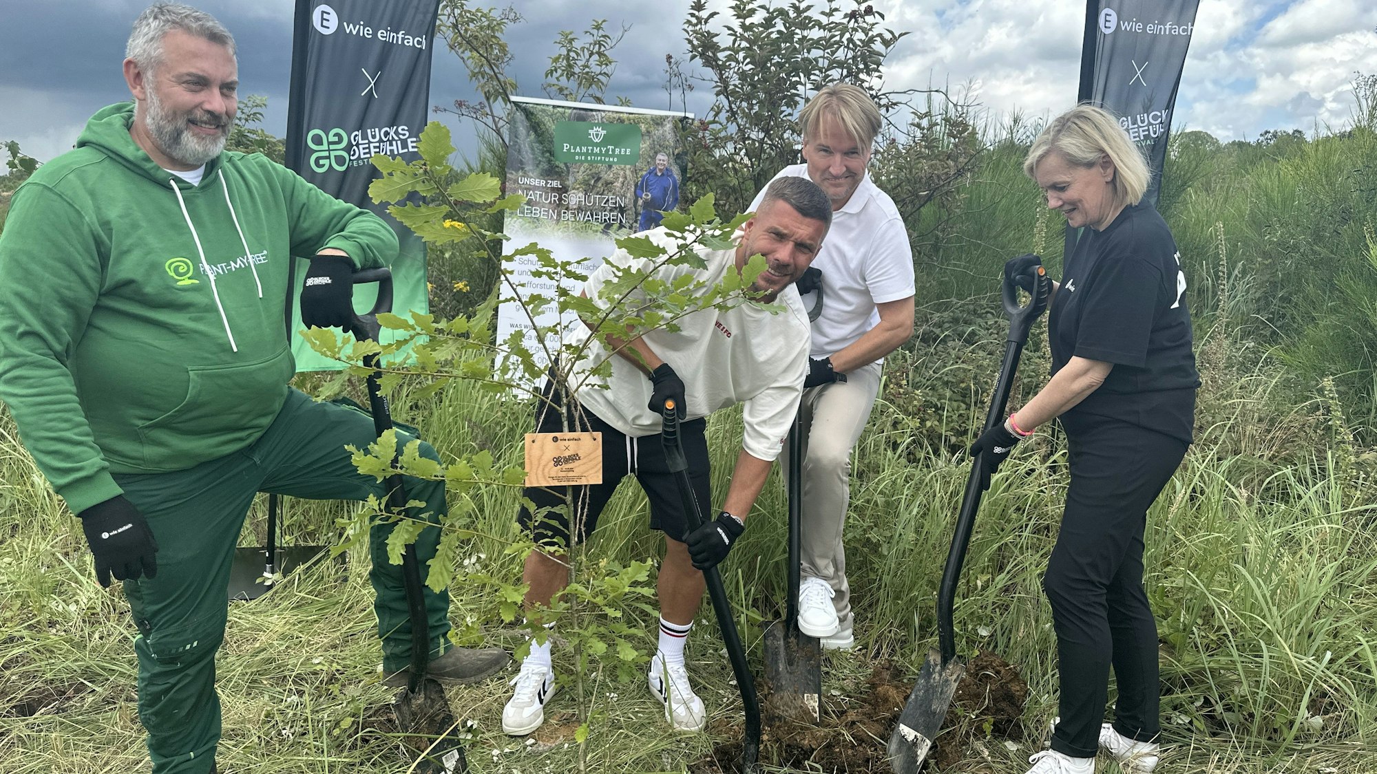 Lukas Podolski und weitere Männer und eine Frau stehen in einem Wald in Lindlar und pflanzen einen Baum.