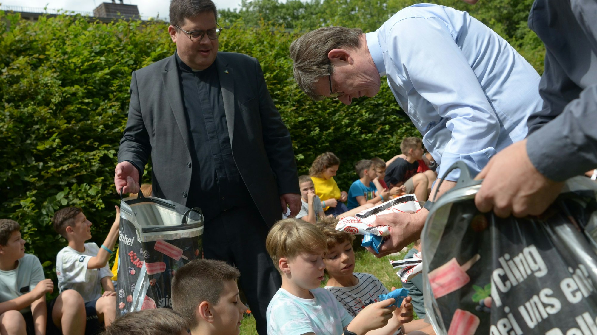 Die Kinder sitzen auf Holzbänken, Woelki reicht ihnen Eis am Stiel.