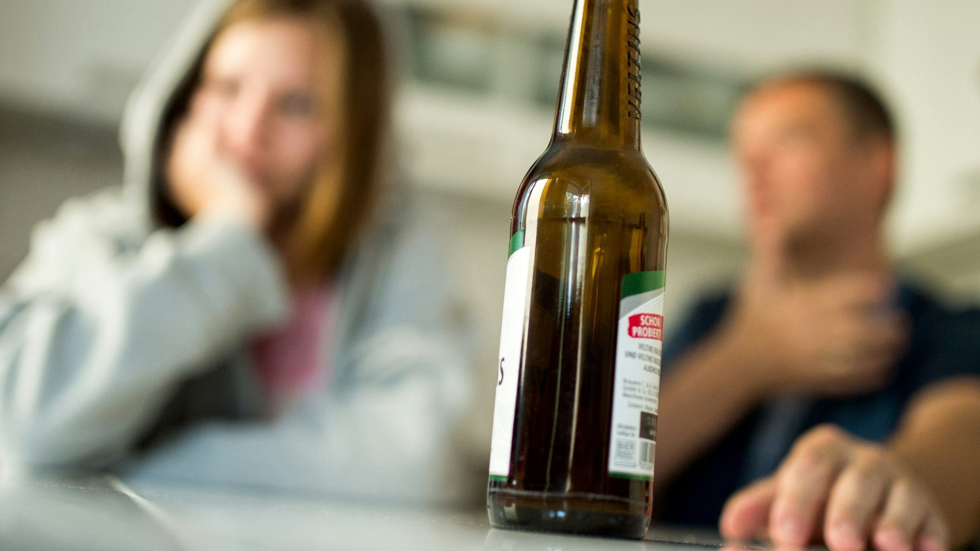 Im Vordergrund steht eine Bierflasche, im Hintergrund sind zwei Personen verschwommen zu sehen. Ein jüngeres Mädchen mit einem grauen Kapuzenpulli und ein Mann im T-Shirt sitzen am Tisch.