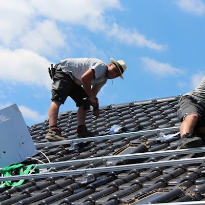 Zwei Dachdecker arbeiten auf einem Dach. Beide tragen Shorts, T-Shirt und Strohhut.
