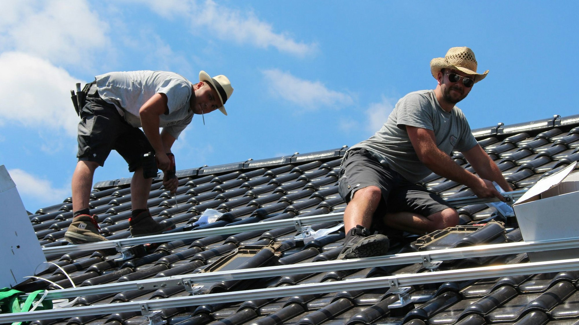 Zwei Dachdecker arbeiten auf einem Dach. Beide tragen Shorts, T-Shirt und Strohhut.