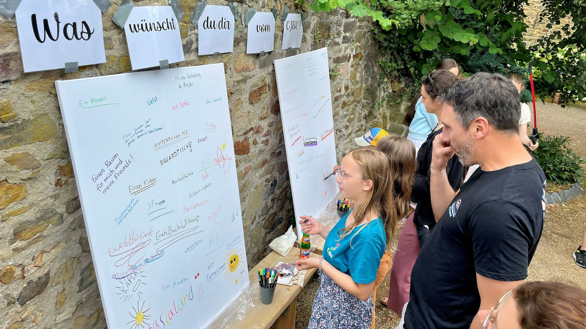 Kinder, denen Erwachsene über die Schulter schauen, stehen vor weißen Tafeln, auf denen schon Wünsche niedergeschrieben worden sind.
