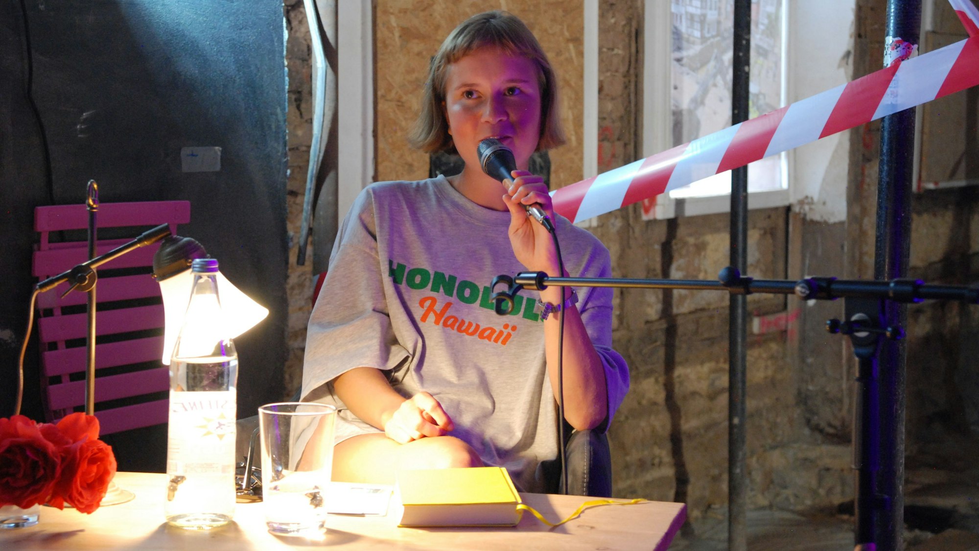 Die Autorin sitzt vor einem Absperrband und spricht in ein Mikrofon. Eine Stehlampe spendet Licht, im Hintergrund eine unverputzte Fachwerkwand.