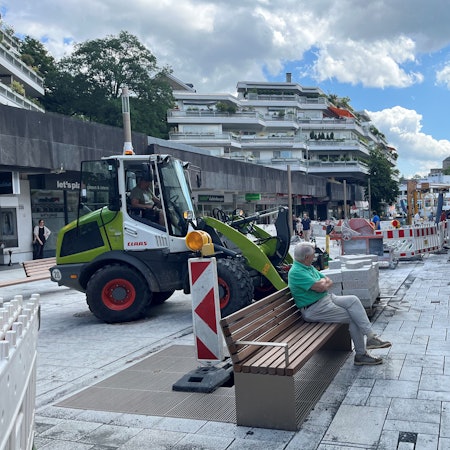 Ein Mann sitzt auf einer Bank, um ihn herum Baustellenabsperrungen, hinter ihm ein grüner Bagger.