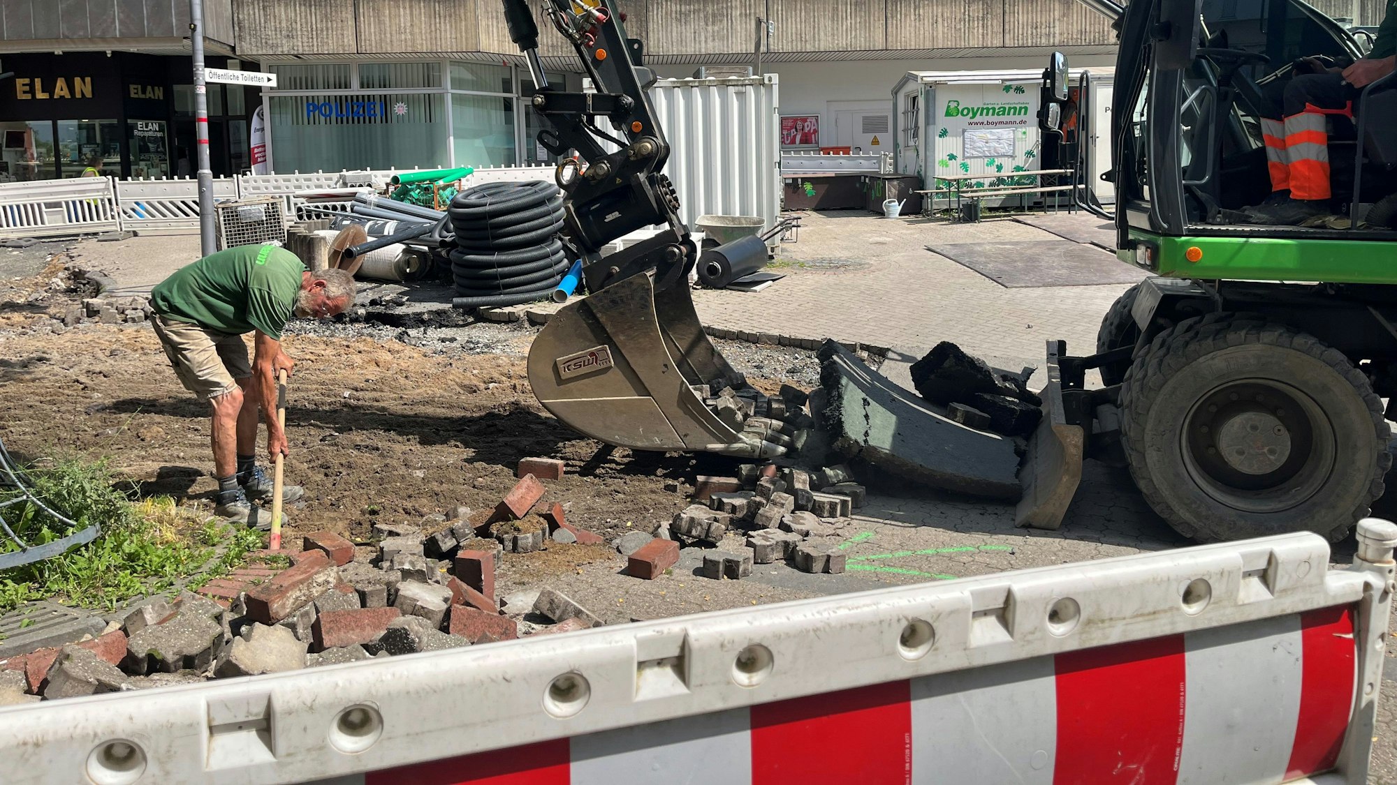 Ein Mann steht mit einer Schaufel zwischen losen Pflastersteinen, vor ihm ein Bagger.