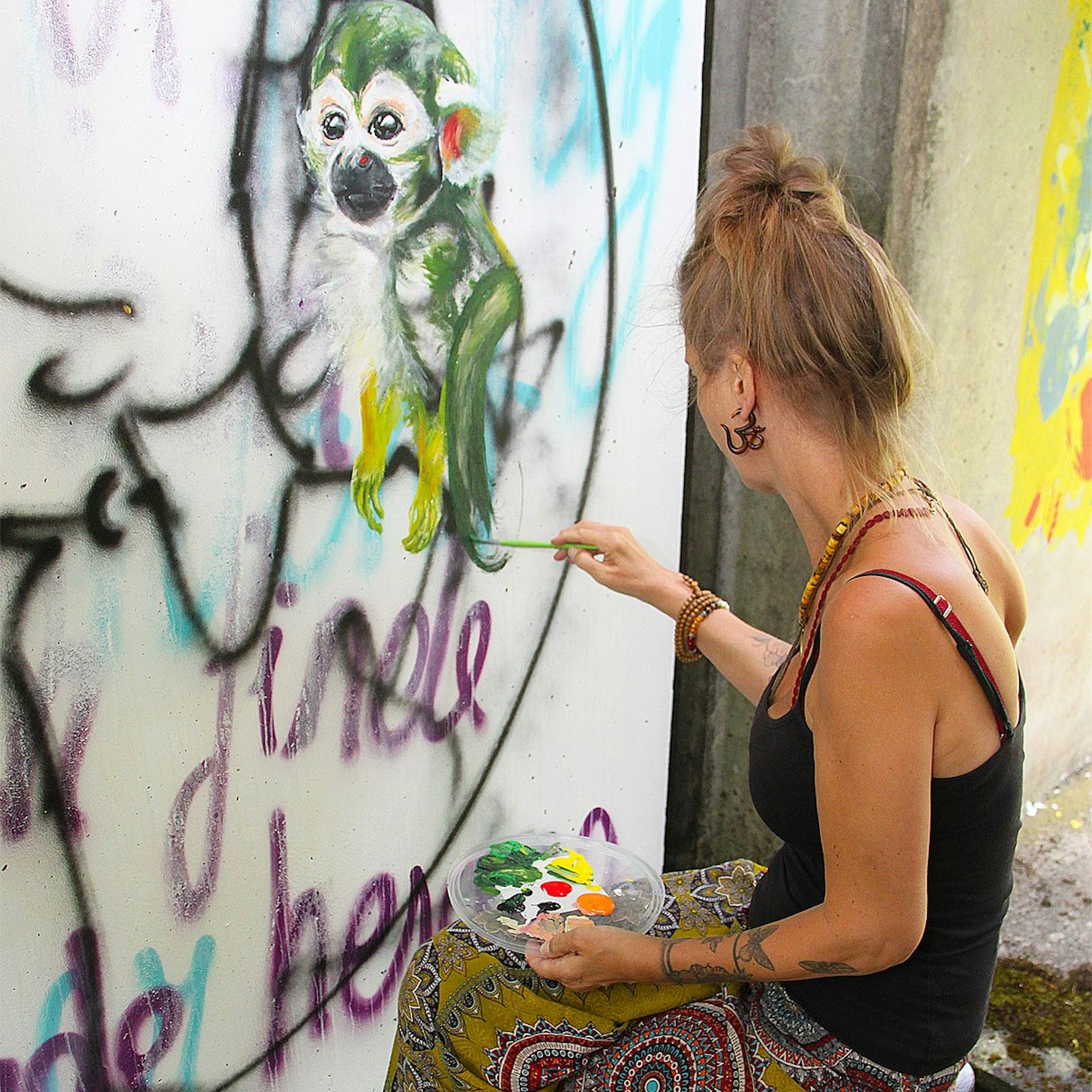 Eine Frau sitzt vor einer Beton-Wand. In der linken Hand hält sie eine Farbpalette. Mit der rechten Hand malt sie in grüner Farbe ein Äffchen.