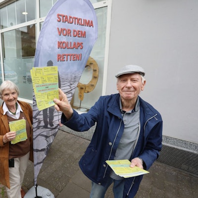 Zwei Männer und eine Frau stehen vor einem Aufsteller mit der Aufschrift „Stadtklima vor dem Kollaps retten" und zeigen Flugblätter.