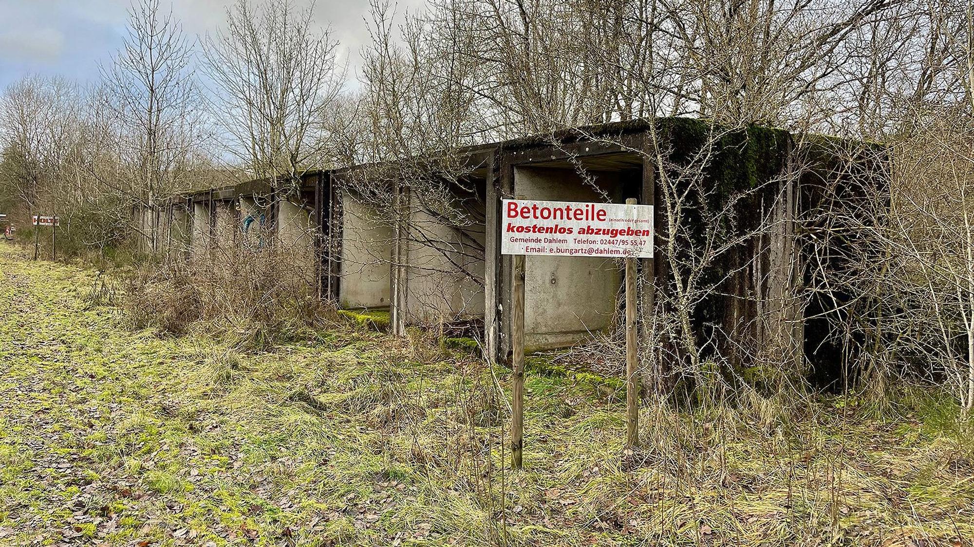 Betonelemente stehen zwischen kahlen Bäumen auf einer Wiese. Auf einem Schild steht: „Betonteile kostenlos abzugeben“.
