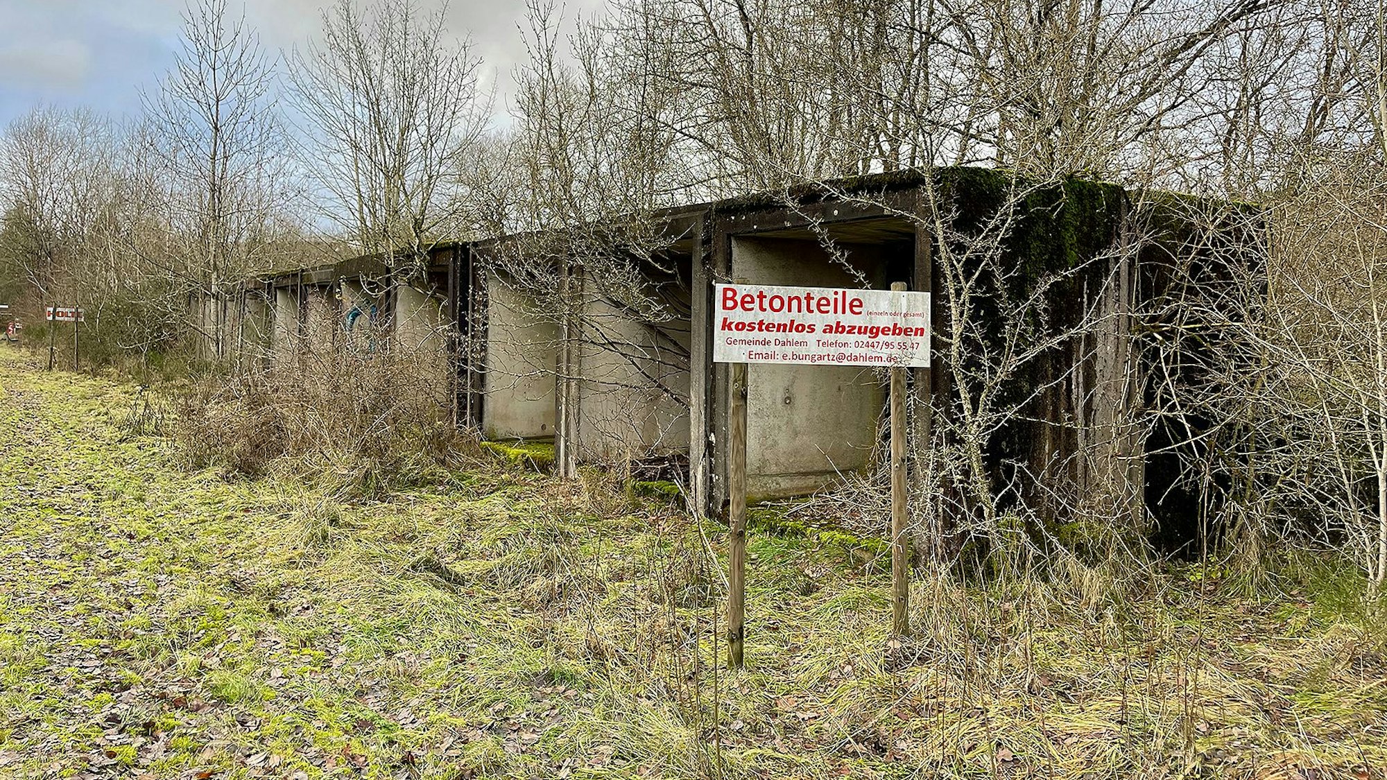 Betonelemente stehen zwischen kahlen Bäumen auf einer Wiese. Auf einem Schild steht: „Betonteile kostenlos abzugeben“.