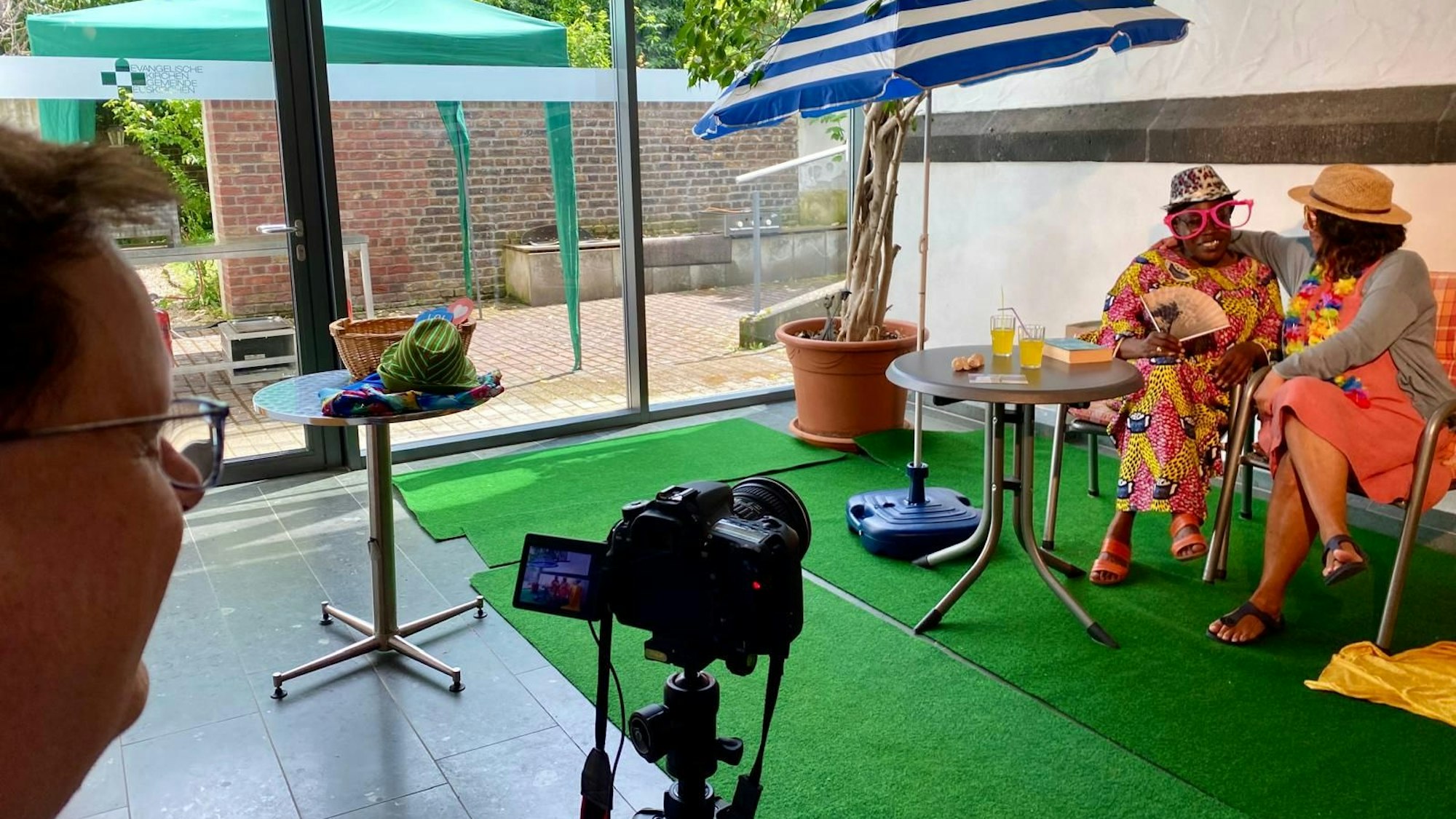 Zwei Frauen tragen Hüte, sitzen unter einem Sonnenschirm und lassen sich fotografieren.