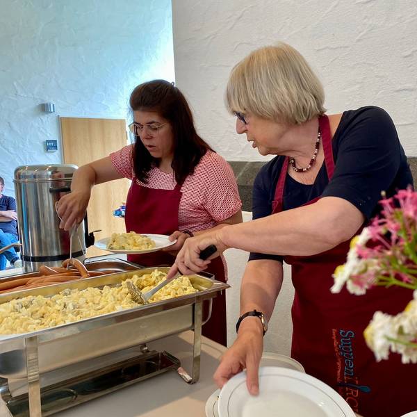 Zwei ehrenamtliche Mitarbeiterinnen verteilen Kartoffelsalat und Würstchen auf Teller.