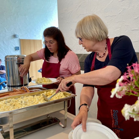 Zwei ehrenamtliche Mitarbeiterinnen verteilen Kartoffelsalat und Würstchen auf Teller.
