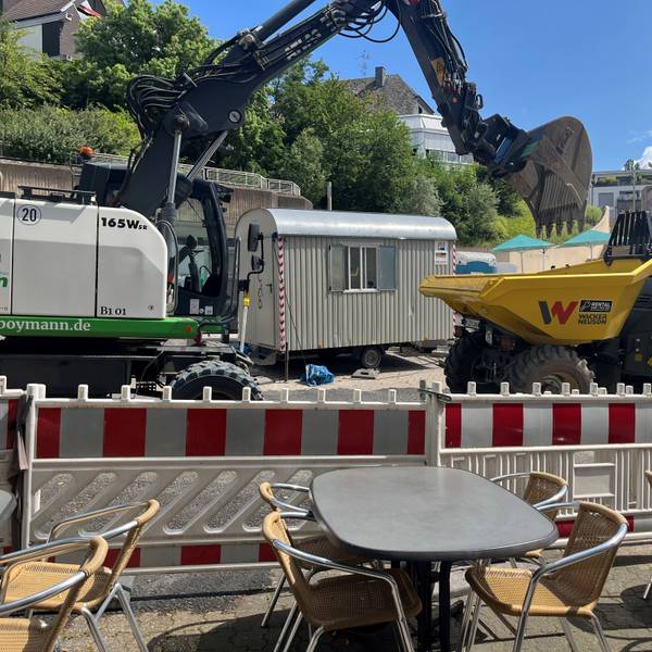 Unmittelbar vor der Absperrung stehen Tische eines Restaurants. Unmittelbar dahinter fährt ein Bagger.