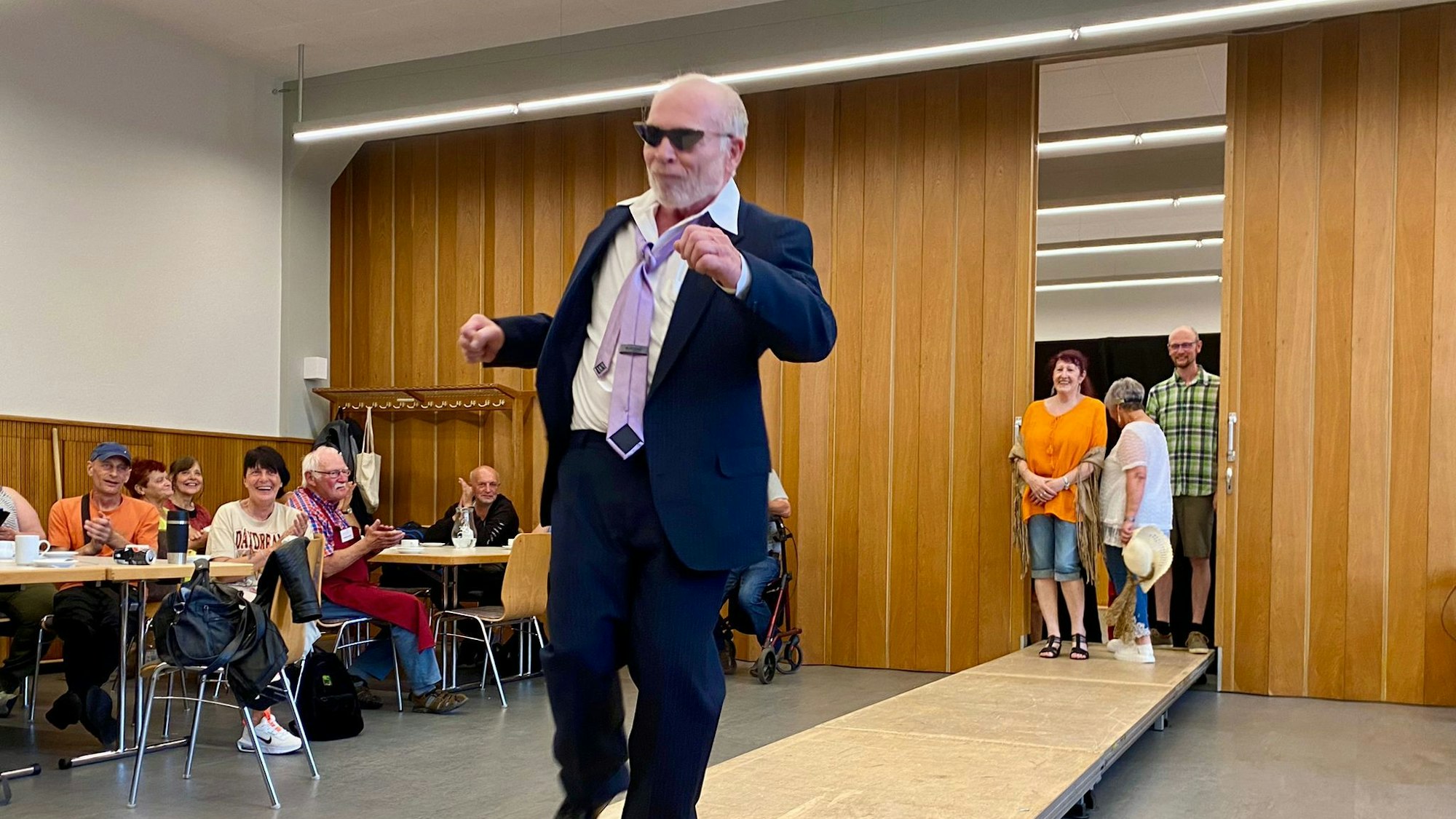 Ein Mann im Anzug tanzt über einen Laufsteg, das Publikum lacht.