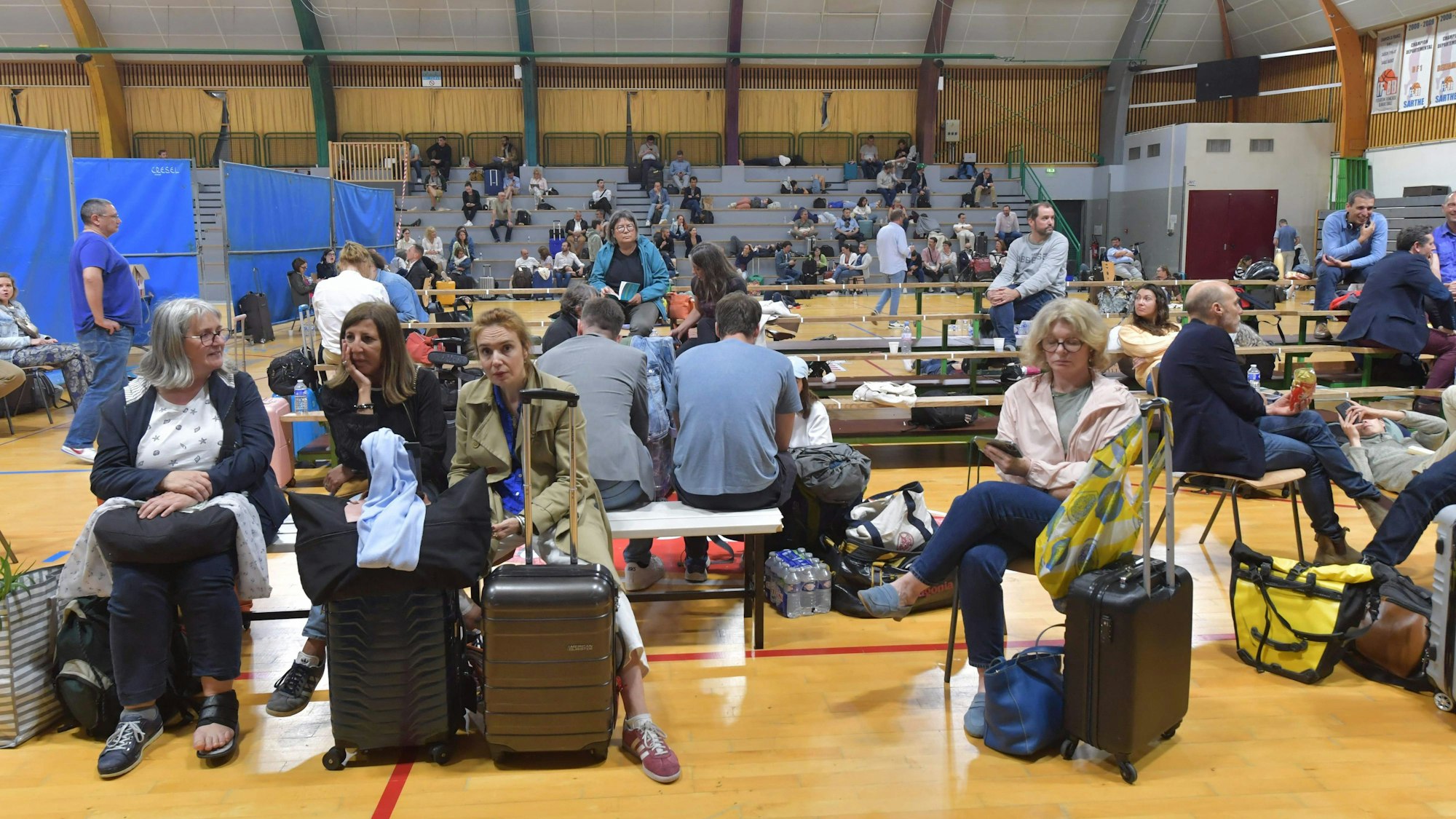 Mehrere Personen sitzen in einer Sporthalle auf Bänken neben ihrem Gepäck.