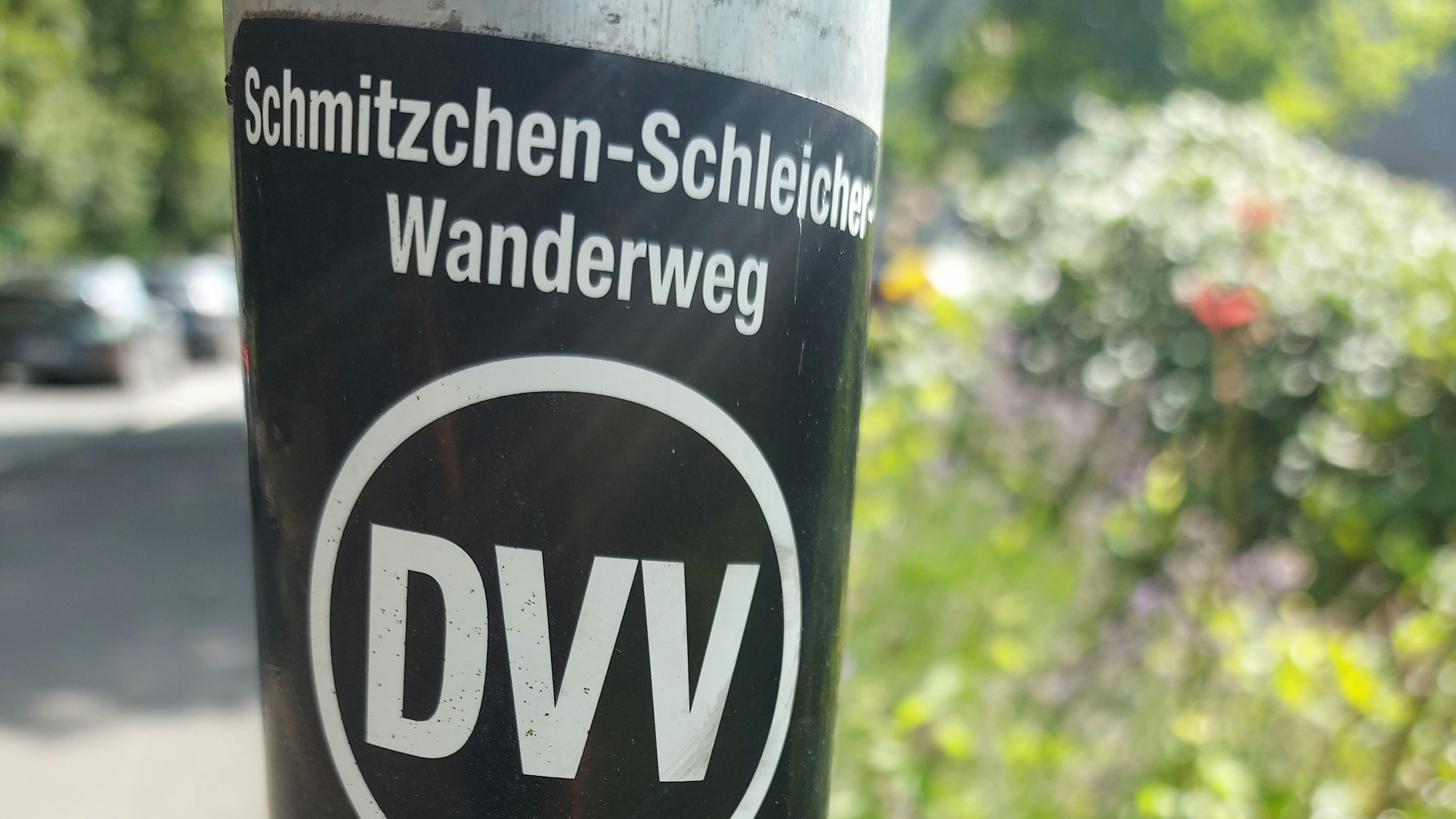 Ein Hinweisschild für den Schmitzchen-Schleicher-Wanderweg, aufgeklebt auf die Stange eines Straßenschildes.