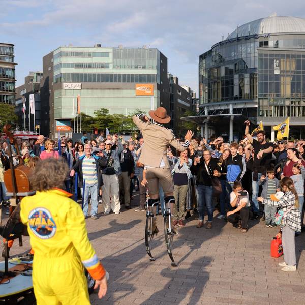 Ein Artist auf Stelzen unterhält eine Menschenmenge auf einem Platz.