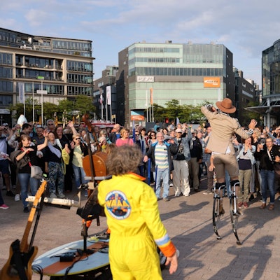 Ein Artist auf Stelzen unterhält eine Menschenmenge auf einem Platz.