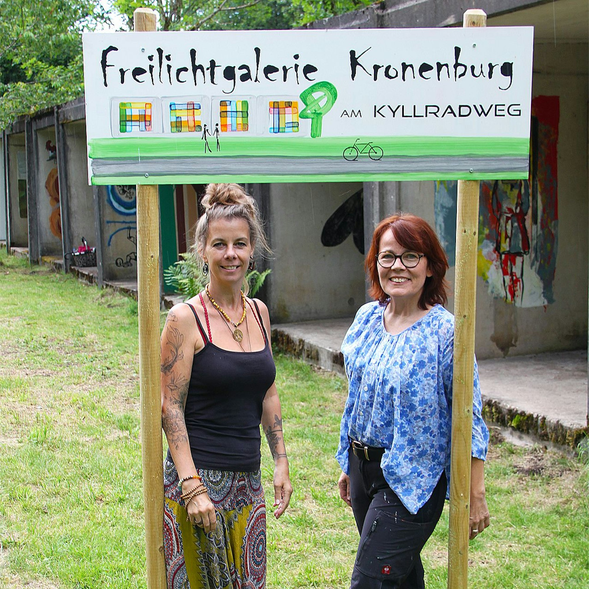Zwei Frauen stehen unter einem Schild, auf dem steht: „Freilichtgalerie Kronenburg am Kyllradweg“.