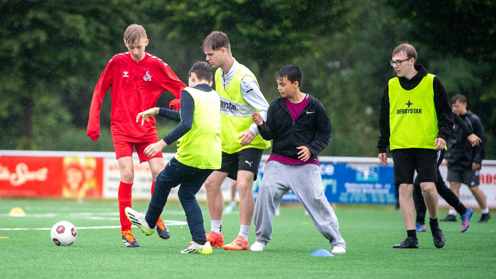 Das Bild zeigt eine Gruppe von Jugendlichen, die auf dem Kunstrasen in Weilerswist im Regen Fußball spielen.