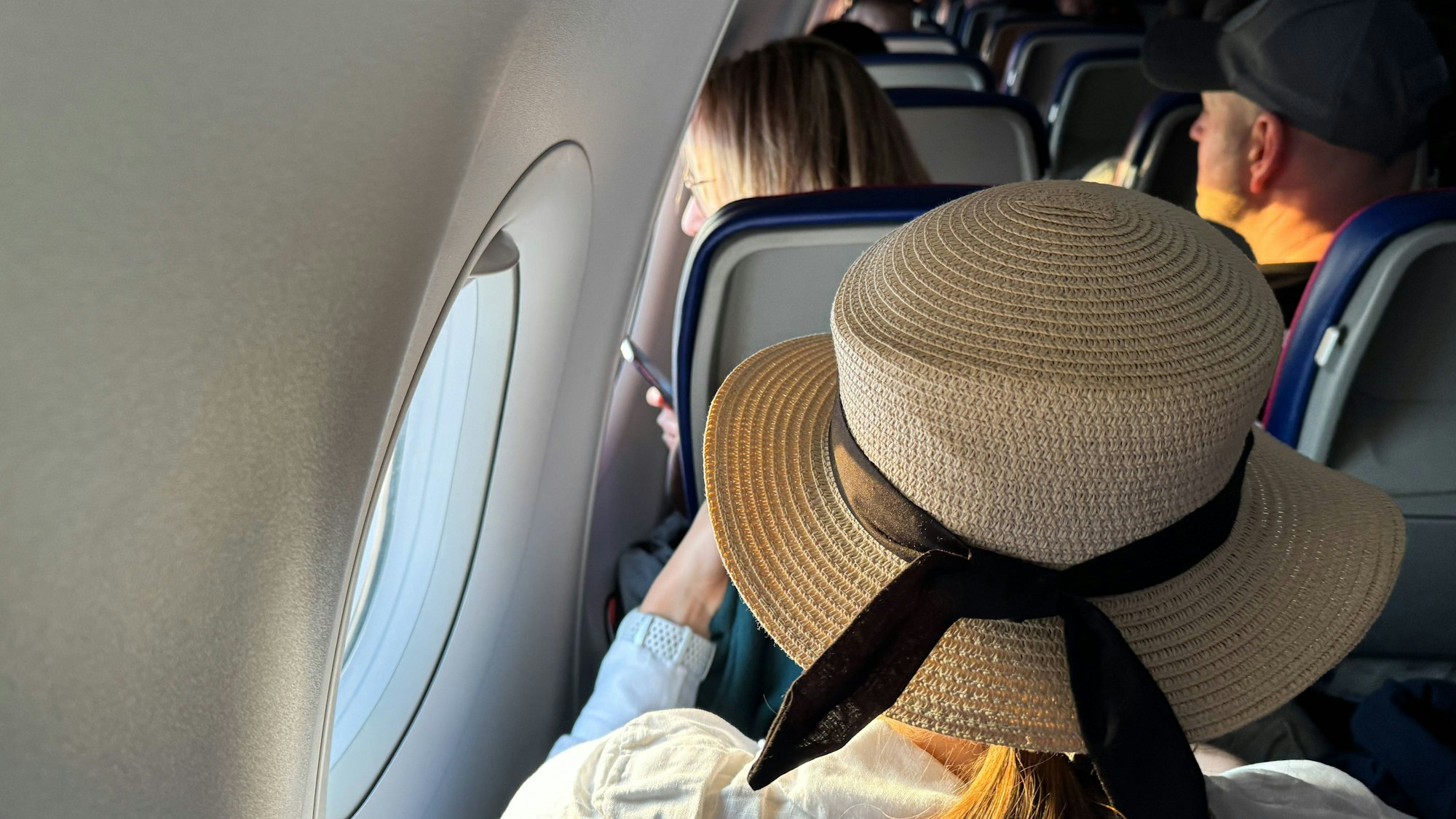 Eine Passagierin sitzt mit Sonnenhut auf einem Fensterplatz im Flugzeug.