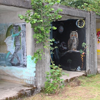 Auf den Wänden von Garagen ähnlichen Beton-Elementen sind Bilder gemalt. Ein Bild zeigt eine rothaarige Frau, ein anderes eine Eule. Um die Betonelemente herum sind Wiese und Bäume.