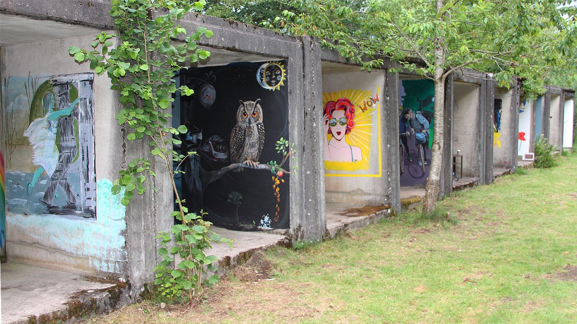 Auf den Wänden von Garagen ähnlichen Beton-Elementen sind Bilder gemalt. Ein Bild zeigt eine rothaarige Frau, ein anderes eine Eule. Um die Betonelemente herum sind Wiese und Bäume.