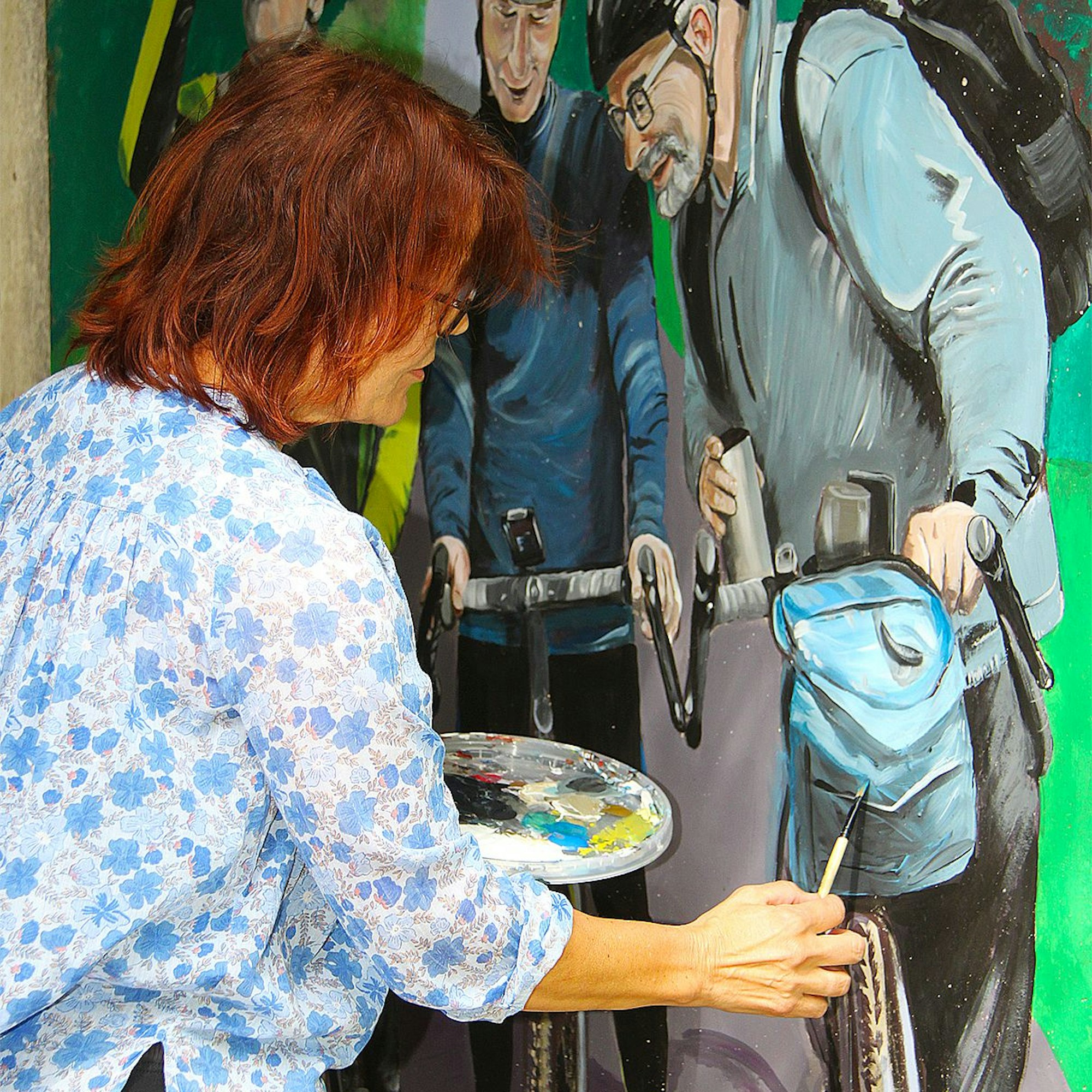 Eine Frau in blau-geblümter Bluse steht mit Farbpalette und Pinsel in der Hand an einer Wand. Sie malt an einem Bild, das drei Radwanderer zeigt.
