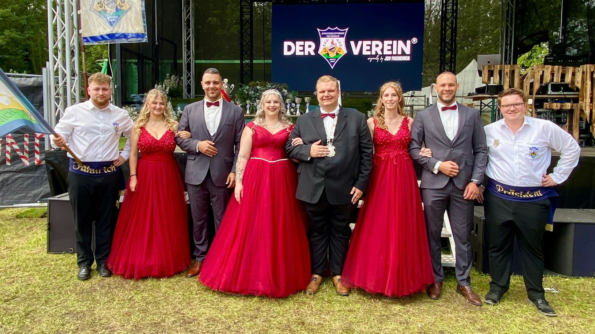 Frauen in roten Kleidern und Männer in Anzügen stehen vor einer Bühne.