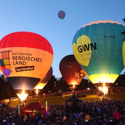 Vier Heißluftballone glühen in der Dämmerung. Tausende sehen zu.