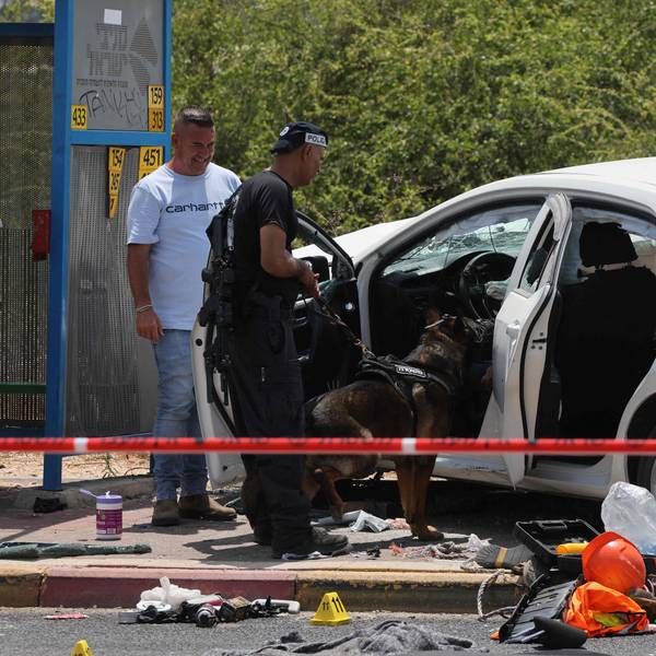 Zwei Männer mit einem Hund stehen an einem beschädigten Auto, das sich hinter einem Absperrband befindet.