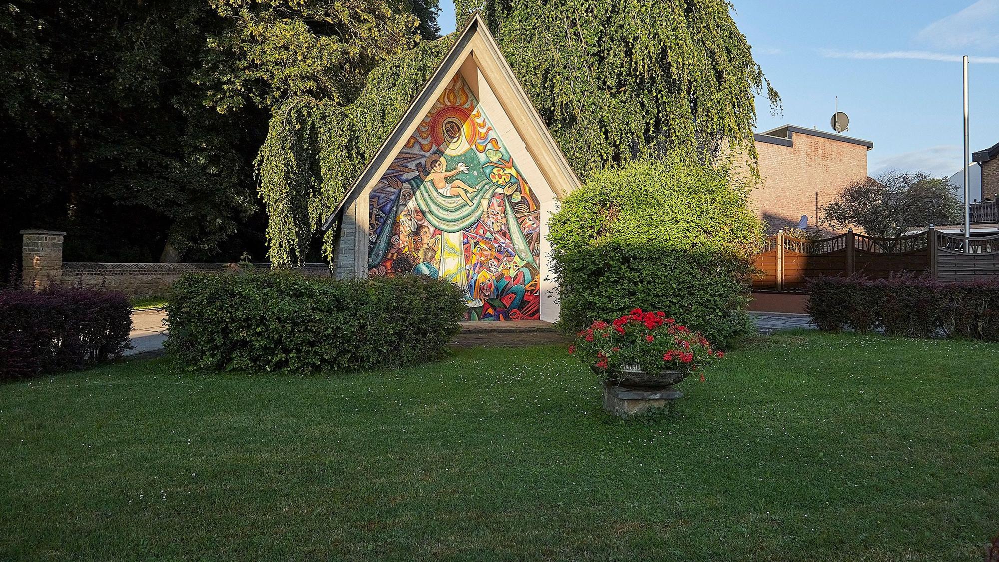 Hinter einer grünen Wiese in Vlatten ist ein Häuschen mit der Darstellung der Schutzmantelmadonna zu sehen.