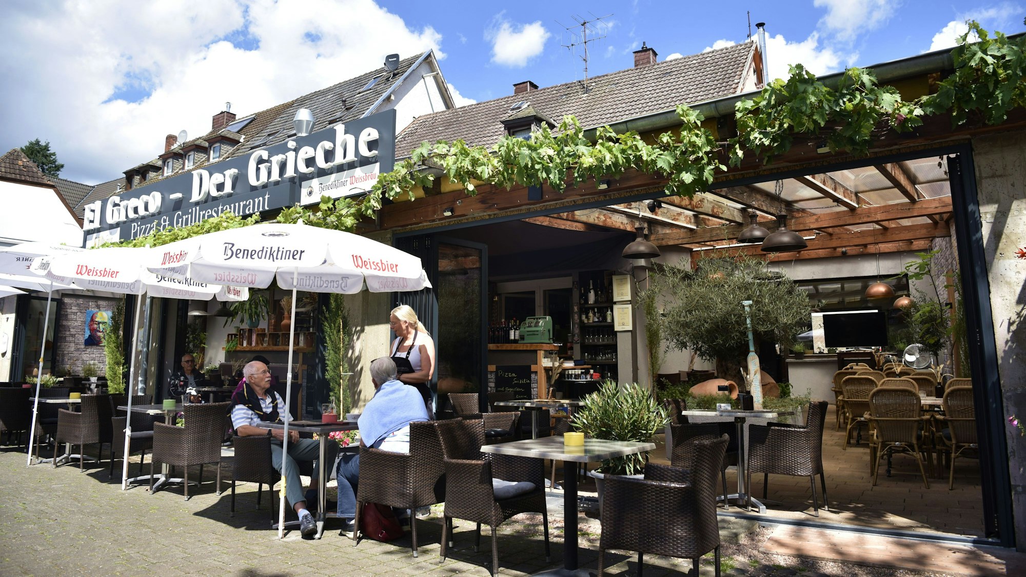 Vor einem Restaurant Stühle und Sonnenschirme, eine Frau mit langen blonden Haaren bedient gerade einen Mann und eine Frau.