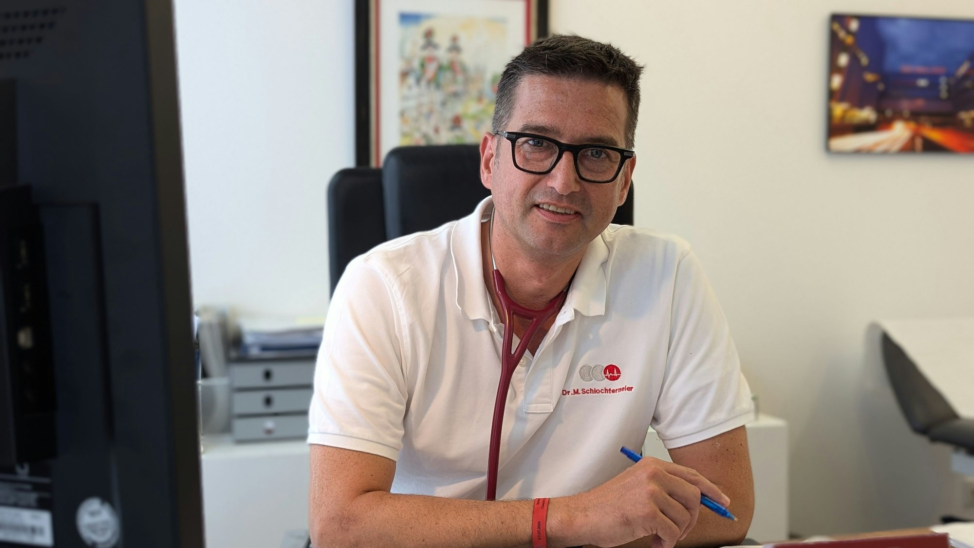 Das Foto zeigt den Arzt im weißen Poloshirt und mit Stethoskop um den Hals und Stift in der Hand an seinem Schreibtisch.