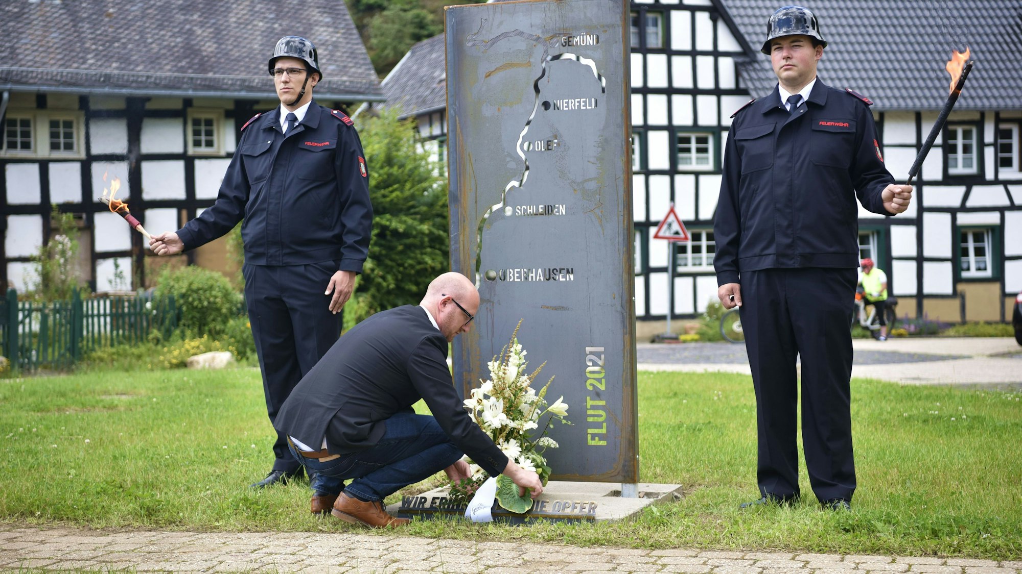 Neben einer Gedenkstele zur Flutkatastrophe 2021 stehen zwei Feuerwehrmänner mit Fackeln. Bürgermeister Ingo Pfennings kniet vor der Stele und legt ein Gesteck ab.