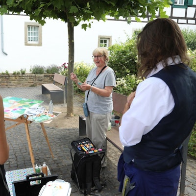 Eine Künstlerin hat eine Staffelei aufgebaut und spricht mit zwei Besuchern.